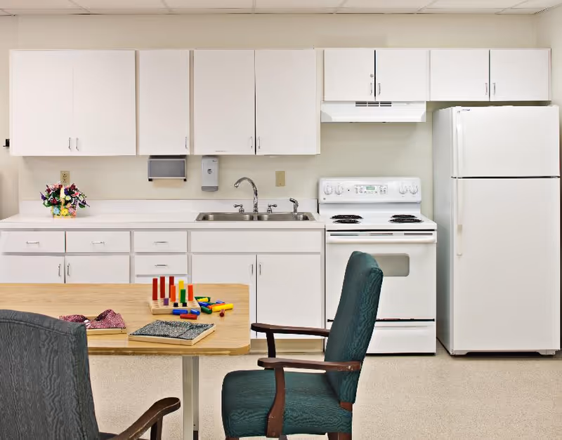 A clean kitchen area with white cabinets, a white stove, a white refrigerator, and a double sink. In front of the kitchen counter is a wooden table with colorful wooden toys and a book on it. Two green cushioned chairs with wooden armrests are positioned around the table.