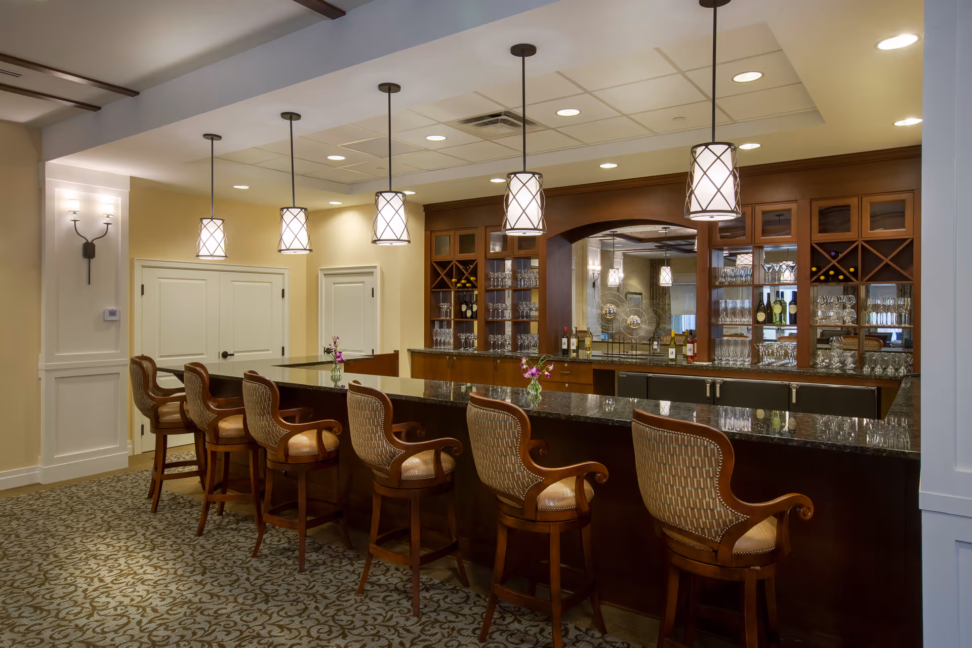 A well-lit bar area with a long granite countertop and six cushioned wooden bar stools. Behind the bar, there are shelves stocked with wine bottles and glassware. Four pendant lights hang from the ceiling above the bar, and the floor is carpeted with a patterned design. The walls are painted in a soft beige color with white trim and paneling.