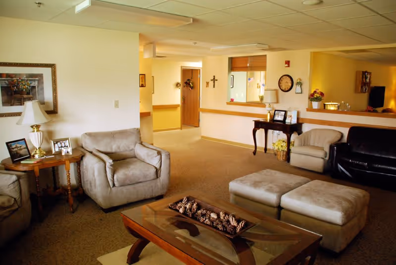 A cozy living room area in a senior care facility with beige armchairs, a black leather sofa, a wooden coffee table with pinecones centerpiece, side tables with lamps and framed photos, and wall decorations including a clock and a cross.