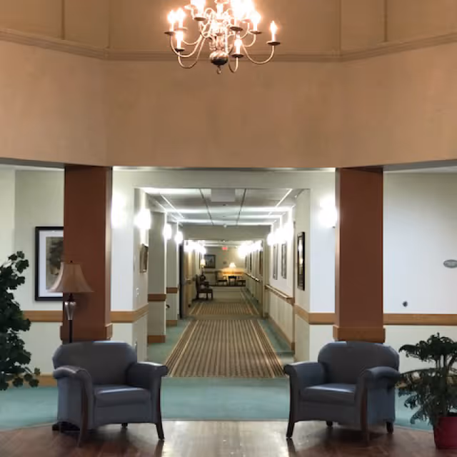 A lobby-style interior showing two armchairs under a chandelier and a long carpeted hallway lined with lights.