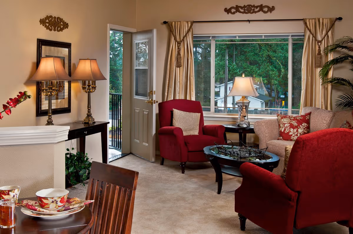 A cozy living room with beige walls and carpet, featuring two red armchairs and a beige sofa with floral cushions. A glass coffee table sits in the center, and a side table with a lamp is placed near a large window with beige curtains. A door is open to an outdoor area, and a console table with two lamps and a mirror is against one wall. A dining table with floral-patterned dishes is partially visible in the foreground.