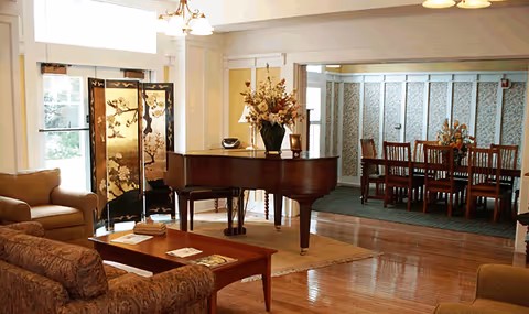 A cozy living room area with a grand piano in the center, a wooden coffee table with magazines, a patterned sofa, and an armchair. In the background, there is a dining area with a wooden dining table and chairs, floral arrangements on the piano and dining table, and a decorative folding screen near the entrance door.