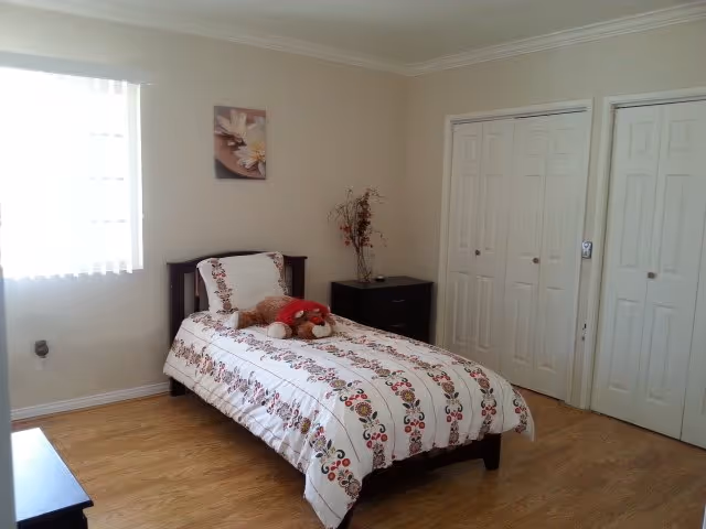 Single bed with floral bedding and a stuffed animal in a bright, simply furnished bedroom with two closets and a window.