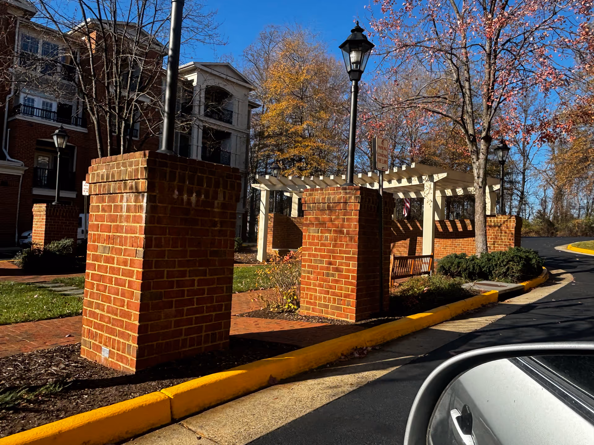 Outdoor view of a senior living facility named Saintsbury Plaza showing brick pillars with lamp posts, a white pergola structure, a wooden bench, trees with autumn leaves, and a curved driveway with a yellow curb. Part of a car's side mirror and door is visible in the foreground.