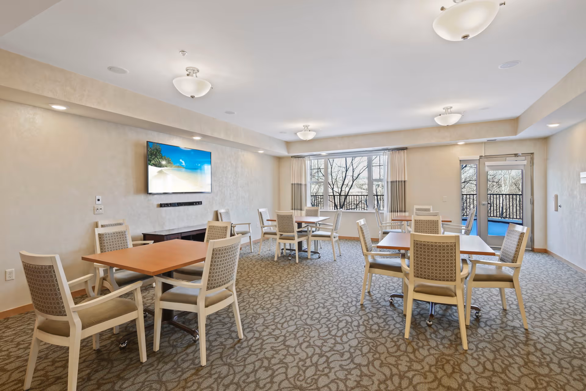 Bright common dining/activity room with multiple tables and upholstered chairs, a wall-mounted TV, and large windows overlooking trees.
