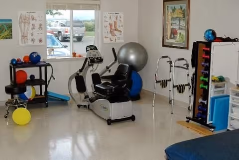 A physical therapy or exercise room with various equipment including a recumbent exercise bike, exercise balls, dumbbells on a rack, walkers, anatomical charts on the walls, and a window showing parked cars outside.