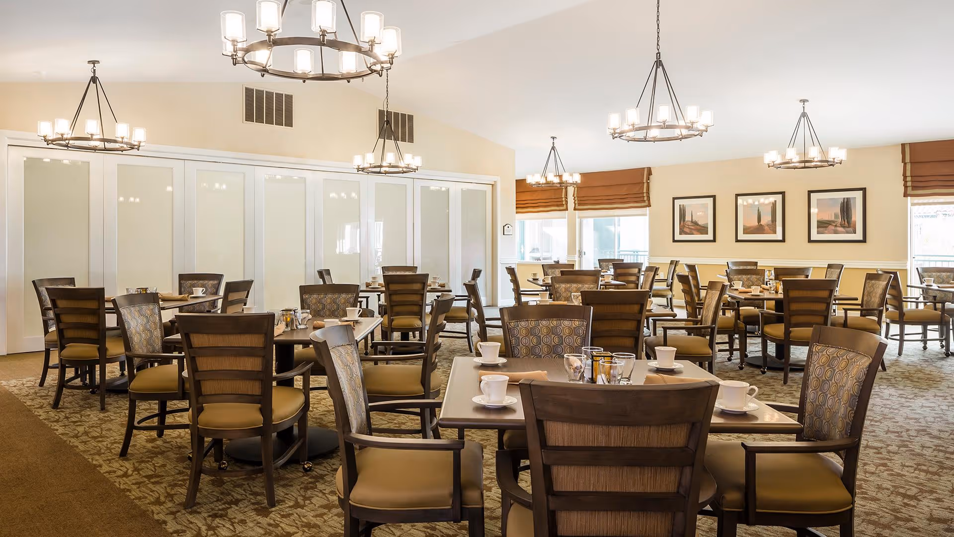 A spacious dining room with multiple wooden tables and chairs arranged neatly. Each table is set with white cups and saucers, napkins, and small condiment containers. The room is well-lit with several modern chandeliers hanging from the ceiling. Large windows with brown Roman shades allow natural light to enter. The walls are decorated with framed landscape artwork.