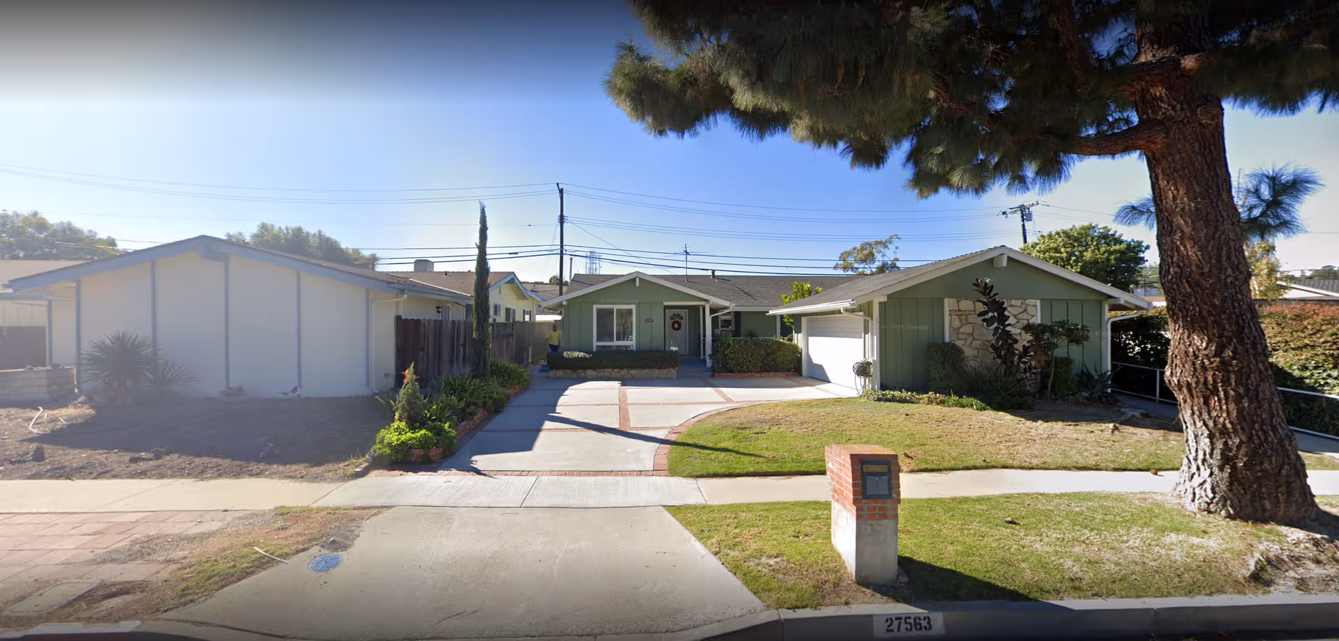 Front exterior view of a single-story residential building with a driveway, garage, and a large tree on the right side. The house has a green and white exterior with some bushes and plants near the entrance.