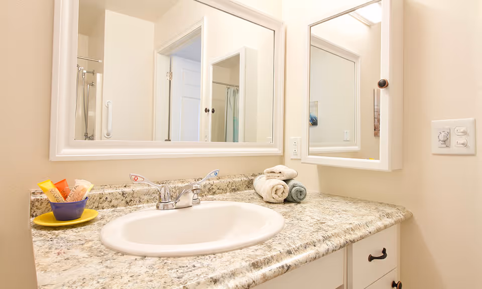 A bathroom countertop with a white sink and silver faucet. On the countertop, there are rolled towels and a small bowl with toiletries. A large mirror and a medicine cabinet with a mirrored door are mounted on the wall above the sink. The bathroom has light-colored walls and a granite countertop.