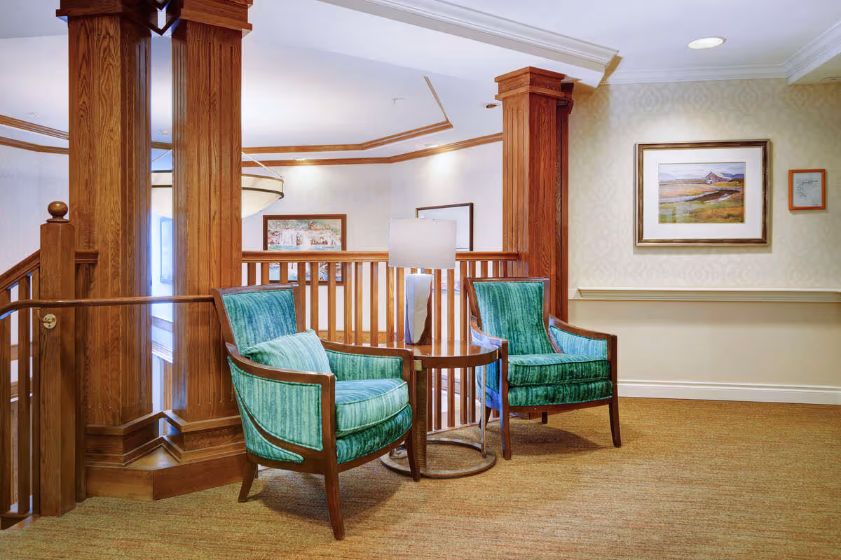 Two green upholstered chairs flanking a small round table with a lamp in a wood-accented interior seating area.