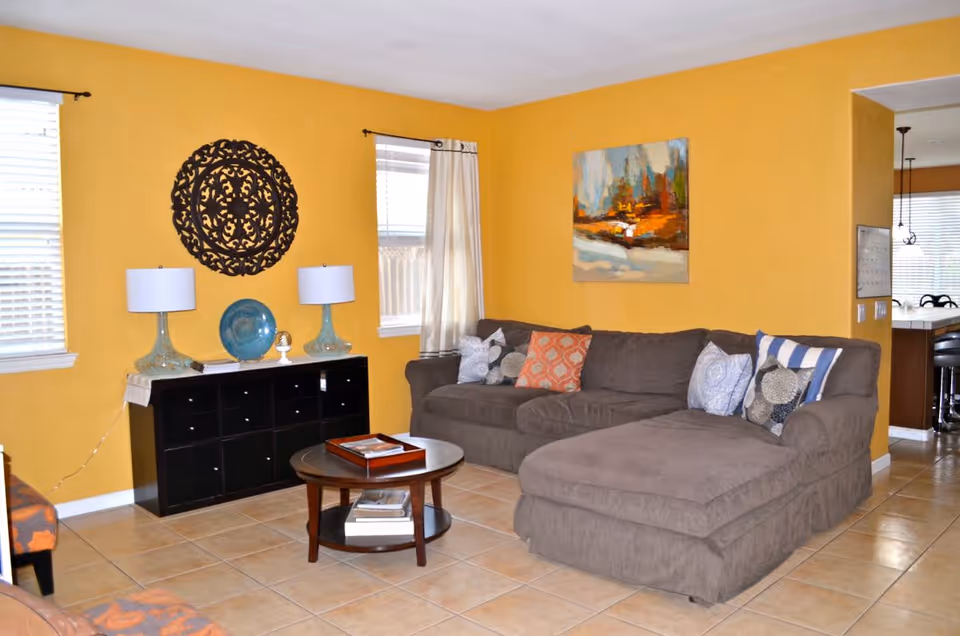 A cozy living room with mustard yellow walls, a large gray sectional sofa with multiple patterned cushions, a round wooden coffee table with books and a tray, a black cabinet with two blue glass lamps and decorative items, two windows with white blinds and curtains, and colorful abstract artwork on the walls.