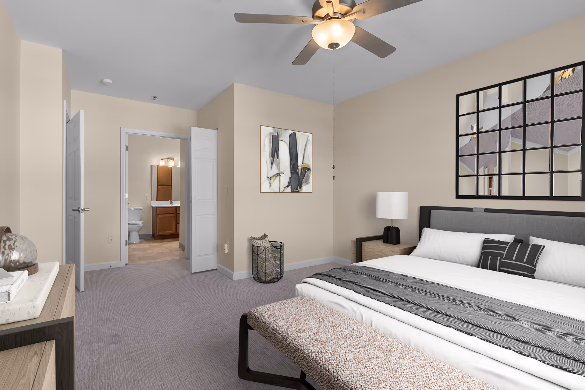 A modern bedroom with a large bed featuring white and gray bedding, a bench at the foot of the bed, a nightstand with a lamp, a decorative wall mirror above the bed, and a ceiling fan. The room has beige walls and carpeted floor. An open door reveals a bathroom with a toilet and sink.