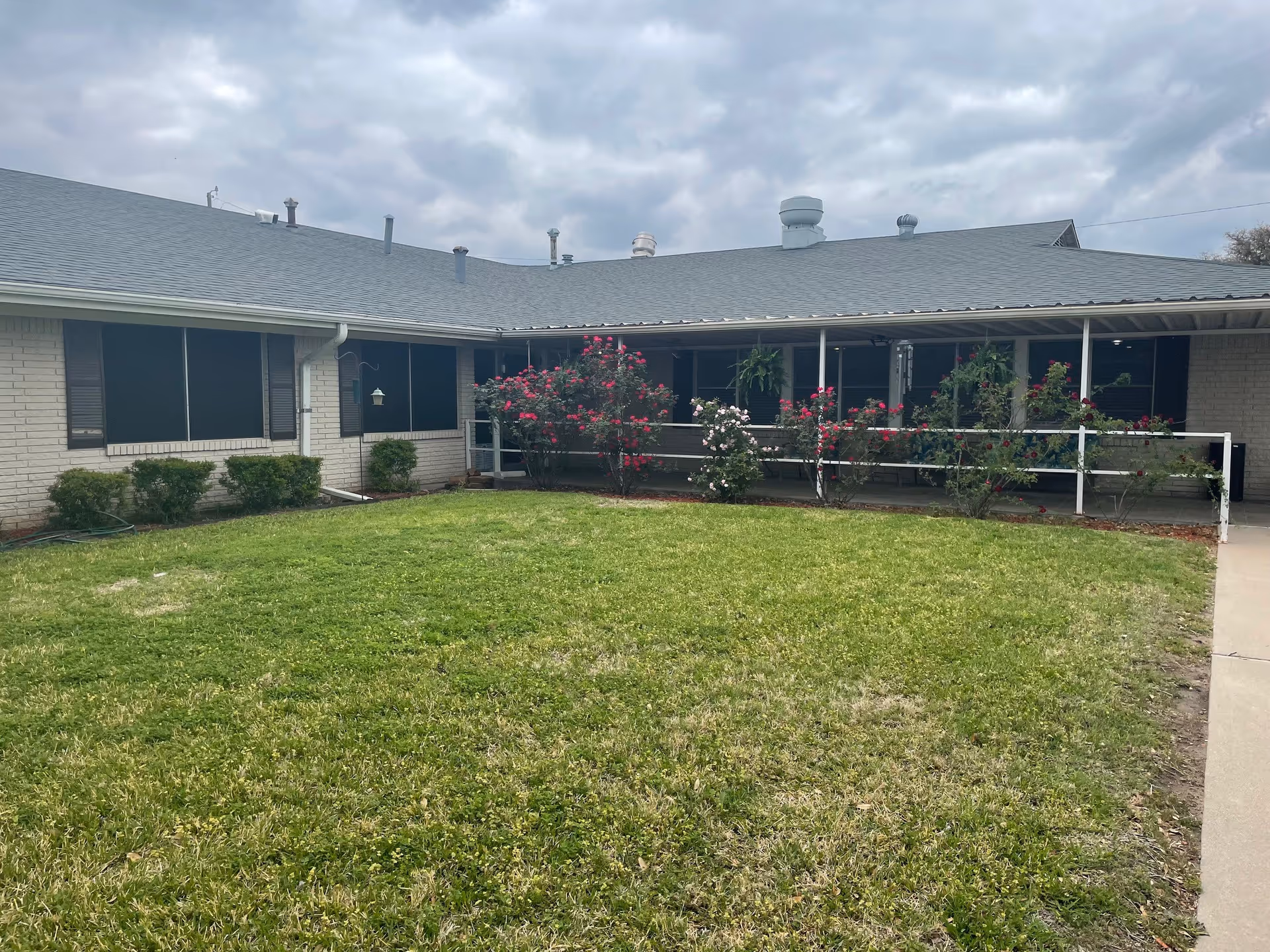 Exterior view of a single-story healthcare residence building with a gray roof, white brick walls, and several windows. There is a green lawn in the foreground with bushes and flowering plants near the building. The sky is cloudy.
