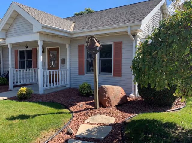 Exterior view of a single-story house with light gray siding and red shutters. There is a small porch with white railings and a wooden door. In front of the house, there is a garden area with red mulch, a large rock, a metal bell mounted on a wooden post, and a stone pathway leading to the porch. Green grass and a leafy tree are visible on the right side under a clear blue sky.