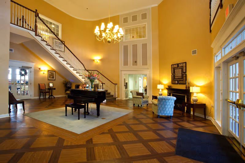 A spacious and elegant lobby area with a grand piano on a light blue rug in the center. The room features a high ceiling with a chandelier, a staircase with wooden railings, comfortable armchairs, side tables with lamps, and large windows letting in natural light. The floor has a patterned wood design, and the walls are painted a warm yellow color.