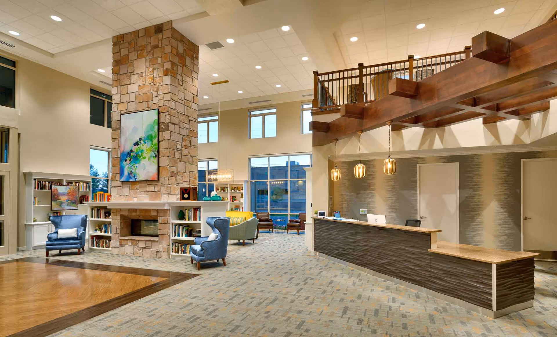Spacious and well-lit senior living facility common area featuring a large stone fireplace with built-in bookshelves, two blue armchairs, a reception desk with pendant lighting, and a mezzanine with wooden railing overlooking the area.