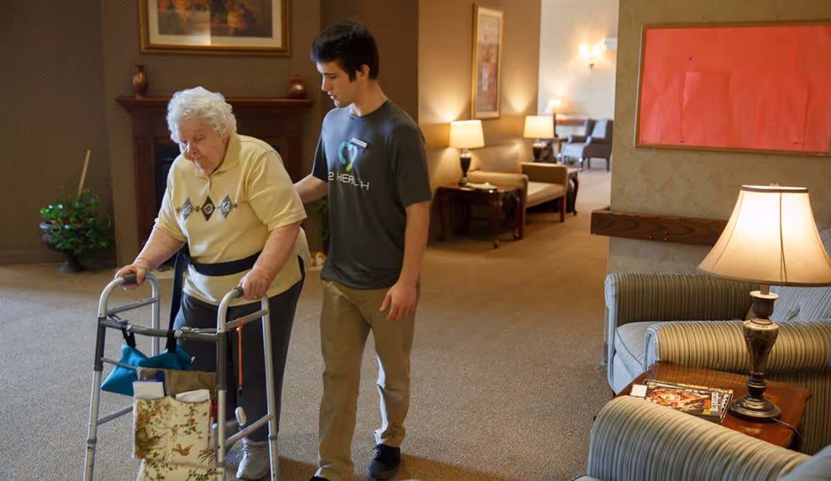 An elderly woman using a walker is being assisted by a young man in a comfortable, well-lit living room area with armchairs, lamps, and framed artwork on the walls.