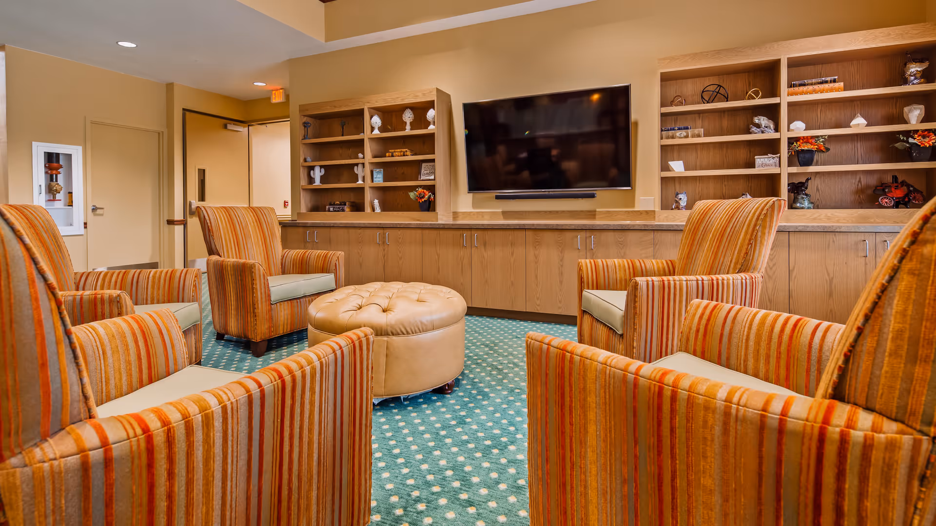 A cozy living room area with five striped orange armchairs arranged around a round beige ottoman. The room features a green carpet with a white dotted pattern, wooden built-in shelves with decorative items, and a large flat-screen TV mounted on the wall.