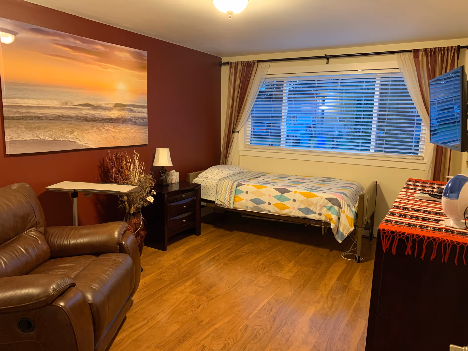 A cozy bedroom with a single bed covered in a colorful geometric patterned blanket, a brown leather recliner chair, a small bedside table with a lamp and decorative items, a large window with blinds and curtains, a wall-mounted TV, and a dresser with a red patterned cloth and a humidifier on top. The walls are painted a warm reddish-brown color with a large sunset beach painting hanging above the recliner.
