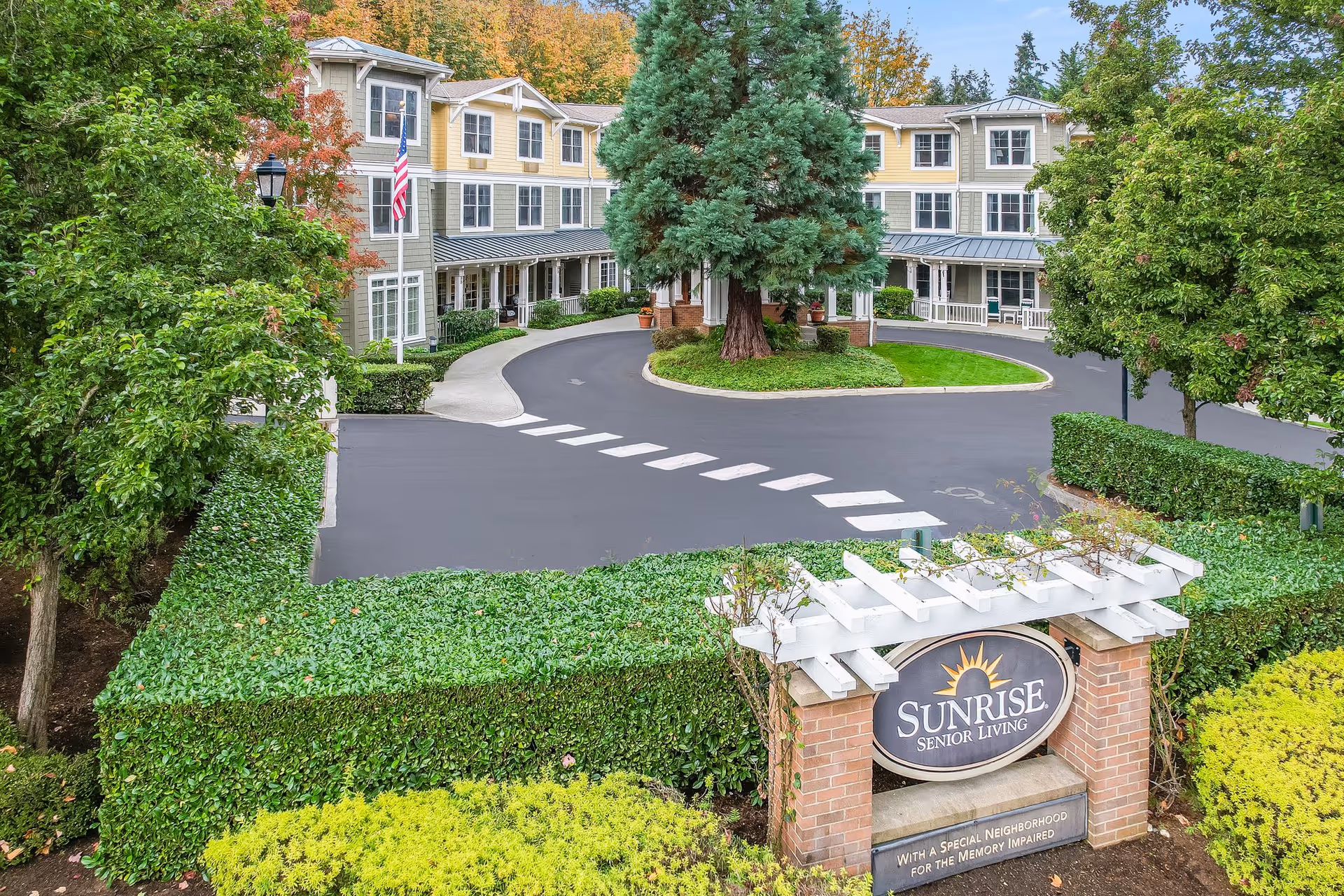 Exterior view of Sunrise Senior Living facility showing a three-story building with yellow and beige siding, surrounded by trees and greenery. A circular driveway with a large tree in the center is visible, along with a sign in the foreground that reads 'Sunrise Senior Living' and mentions a special neighborhood for the memory impaired.