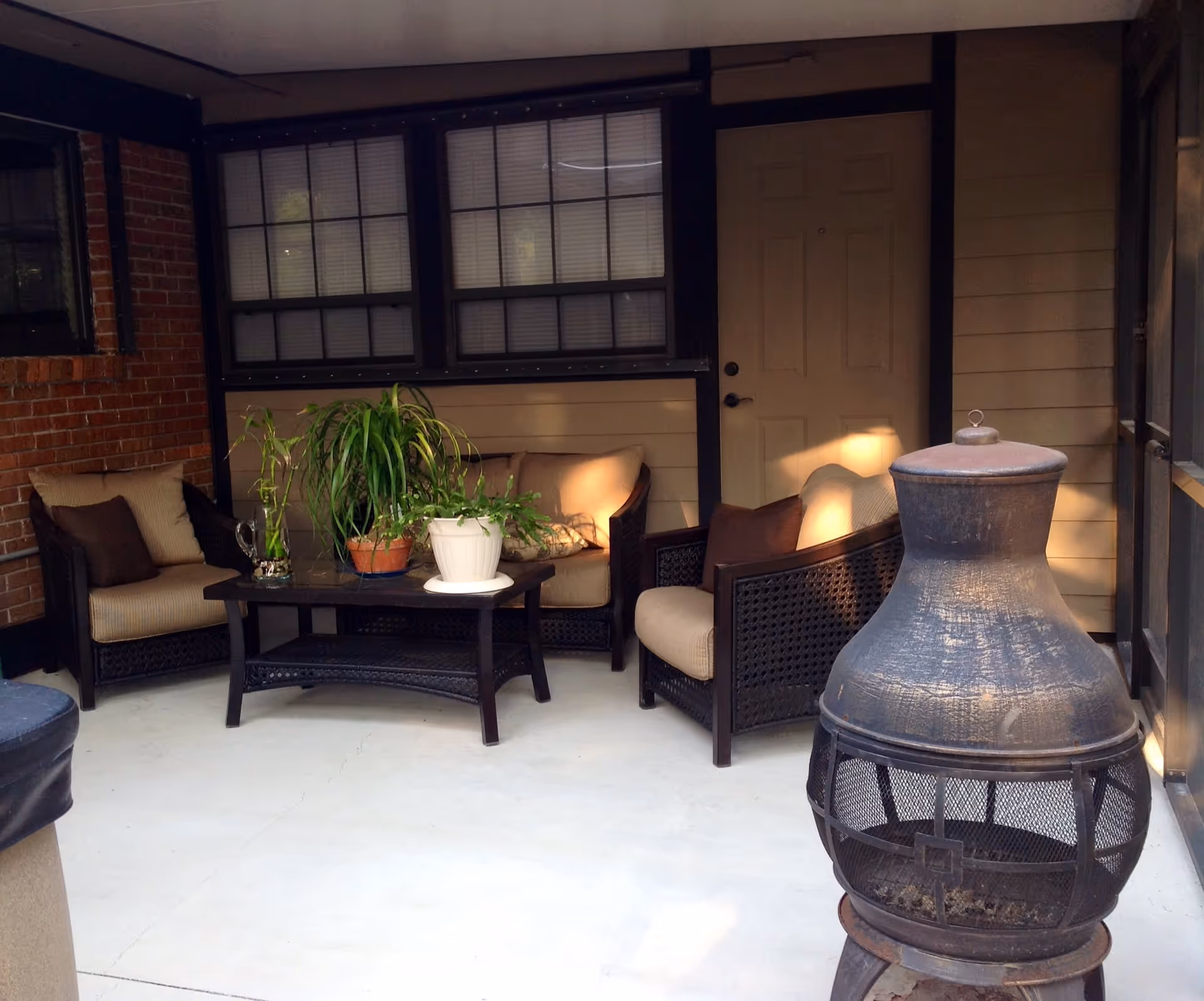A cozy outdoor patio area with cushioned wicker chairs and a matching coffee table holding potted plants. The patio is enclosed with windows and a door, and there is a large metal chiminea fireplace on the right side.