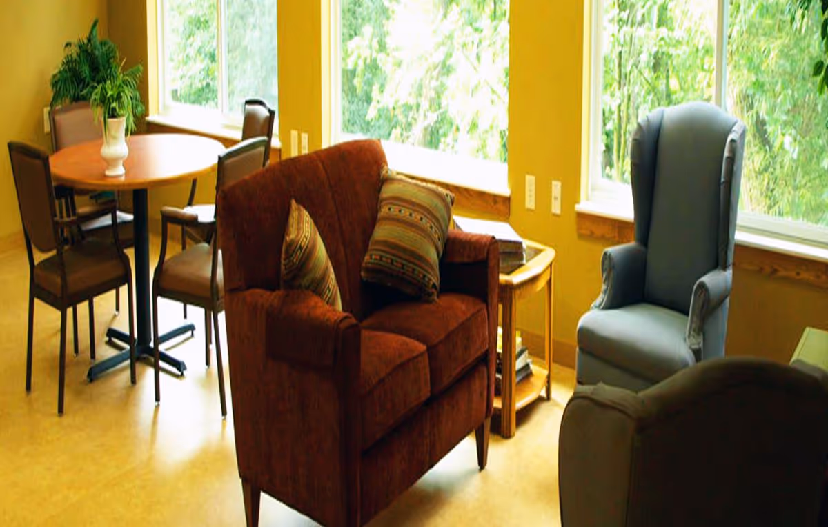 Sunlit common room with upholstered chairs, a loveseat, side table and a round dining table by large windows.