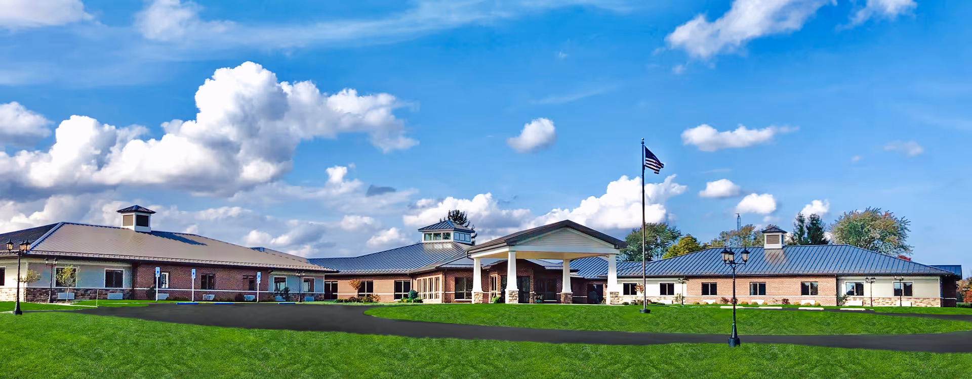 Wide exterior view of The Legacy Assisted Living Community building under a blue sky with scattered clouds. The building has a low, sprawling design with a metal roof, brick and stone facade, and a covered entrance supported by columns. A flagpole with an American flag and several lamp posts are visible on the well-maintained green lawn in front.
