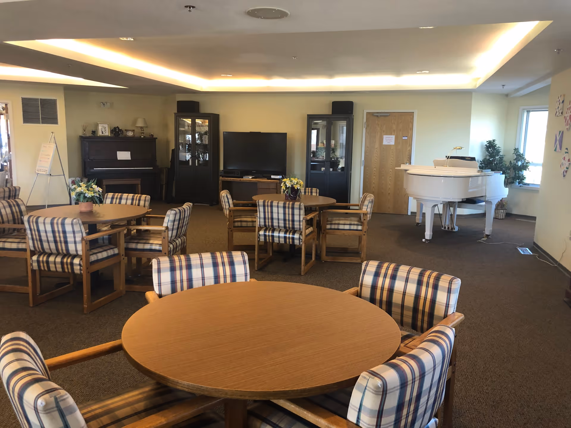 A senior living facility common room with round wooden tables surrounded by cushioned chairs with plaid upholstery. In the background, there is a white grand piano near a window, a dark upright piano, two glass-front cabinets, and a flat-screen TV on a wooden stand. The room is carpeted and has soft ceiling lighting.