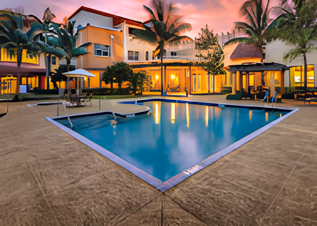 A lit outdoor swimming pool in a courtyard surrounded by a multi-story building and palm trees at dusk.