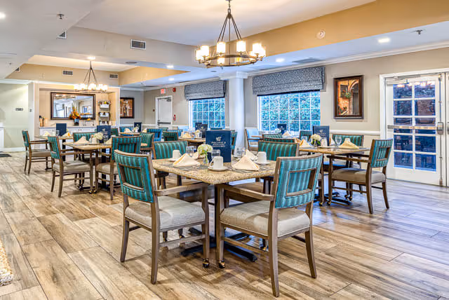 A bright and spacious dining room in a senior living facility with multiple tables set for meals. Each table has neatly arranged napkins, menus, and small flower vases. The room features wooden flooring, large windows with blinds, and modern chandeliers hanging from the ceiling. There are framed artworks on the walls and a glass door leading outside.