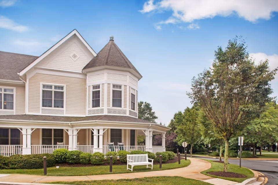Exterior view of a beige two-story building with white trim and a wraparound porch featuring rocking chairs. There is a white bench on the lawn near a curved sidewalk, surrounded by green bushes and trees under a partly cloudy blue sky.