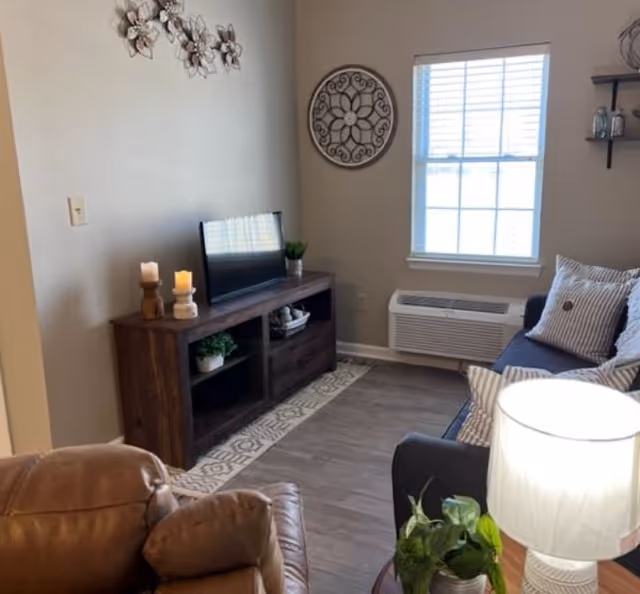 A cozy living room with a brown leather recliner, a dark wooden TV stand holding a flat-screen TV and decorative items, a gray couch with striped pillows, a window with blinds, a wall-mounted air conditioning unit, a round wall decoration, and a lit table lamp on a side table with a small plant.