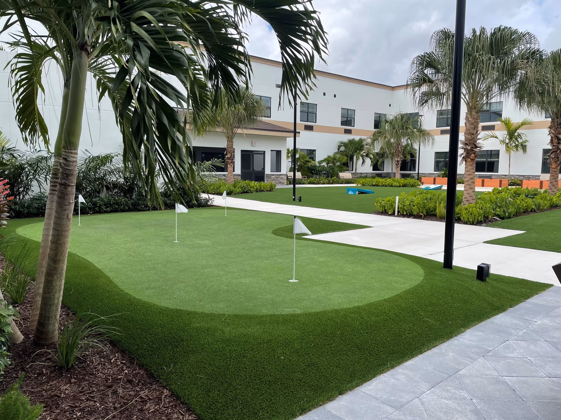 Outdoor courtyard area at Bella Mar Royal Palm Beach featuring a putting green with several small flags, palm trees, landscaped plants, benches, and a cornhole game set on artificial turf, surrounded by a two-story building under a cloudy sky.