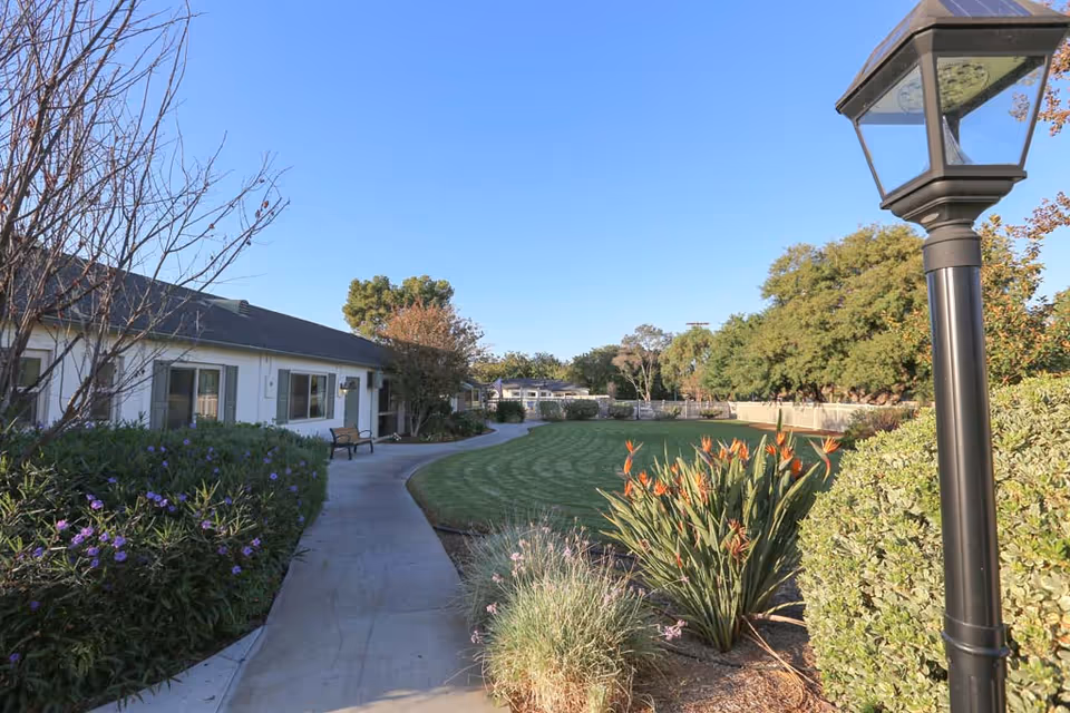 Curved concrete path through a landscaped courtyard with flowering plants, a bench, lamp post, and single-story senior living building under a clear blue sky.