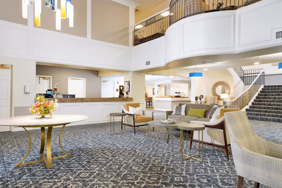 A spacious and well-lit senior living facility lobby with patterned carpet, a round table with a floral arrangement, comfortable seating including armchairs and a sofa, a reception desk in the background, and a staircase leading to an upper floor.