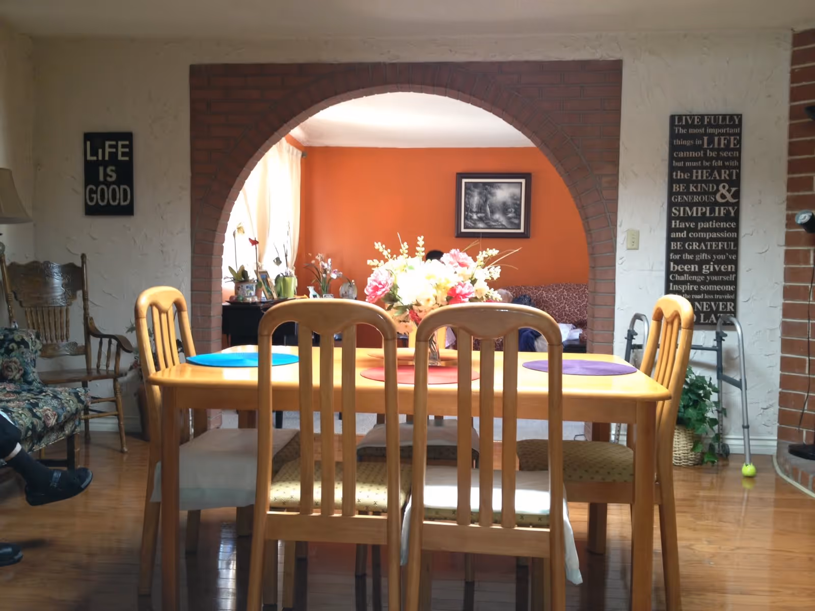 A cozy dining room with a wooden table and six chairs, each with a colorful placemat. A floral centerpiece decorates the table. The room features an arched brick doorway leading to a living area with an orange accent wall, a framed picture, and a sofa. On the left wall, there is a sign that reads 'LIFE IS GOOD,' and on the right wall, a larger sign with an inspirational quote. A walker and a plant are also visible near the right wall.