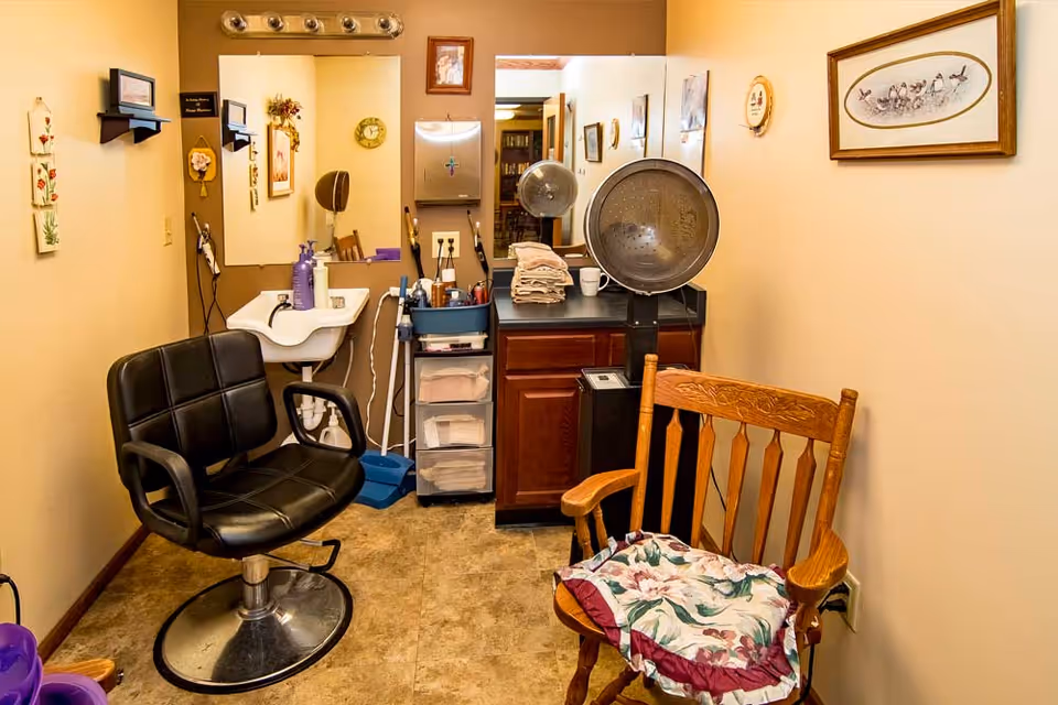 A small hair salon room with a black salon chair, a wooden chair with a floral cushion, a sink with hair care products, a hair dryer hood, and a mirror on the wall. The walls are decorated with framed pictures and small shelves.