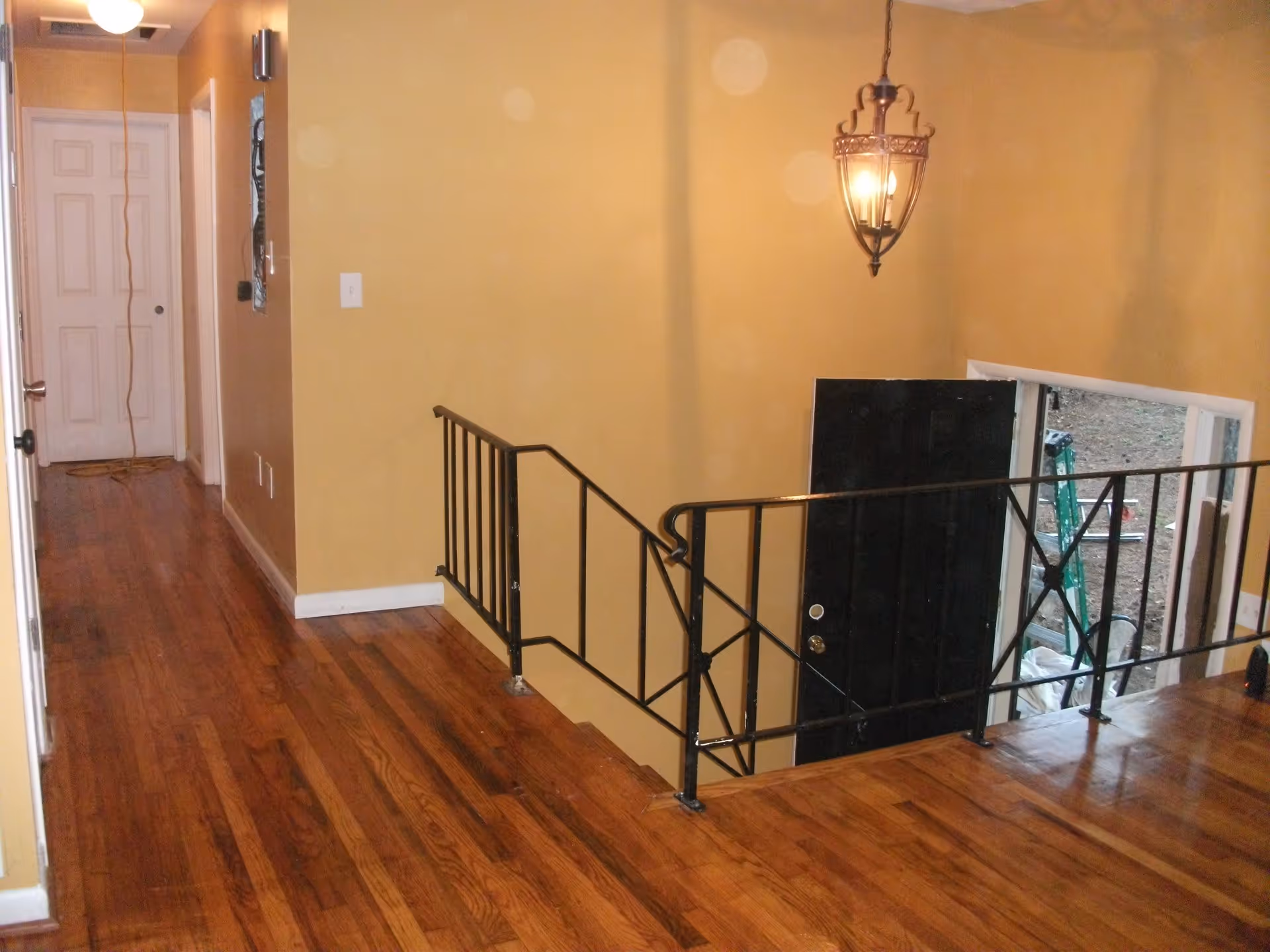 Interior view of a hallway with wooden flooring and yellow walls. There is a black metal railing along the edge of a staircase leading down to a black door that is open to the outside. A hanging light fixture is visible above the staircase.