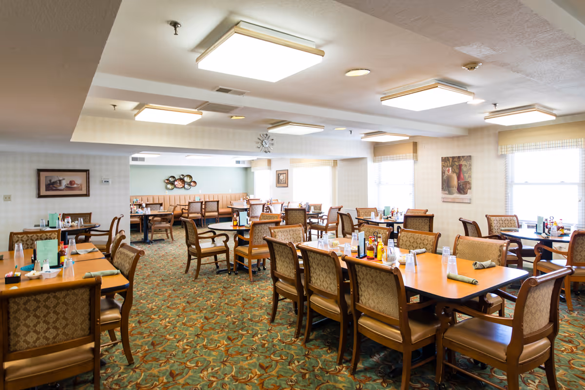 Spacious dining room with multiple tables and chairs set for meals in an assisted living facility.