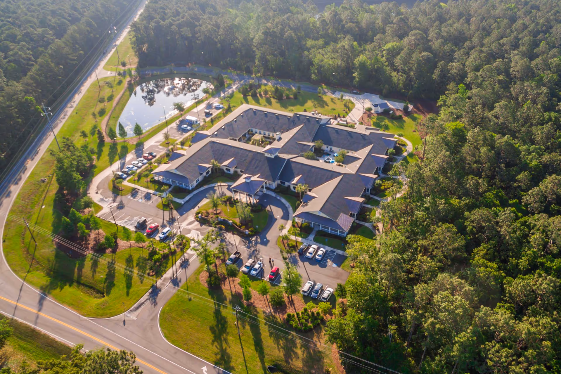 Aerial view of a single-story senior living facility with a central courtyard, surrounding parking lots, a pond, and wooded landscaping.
