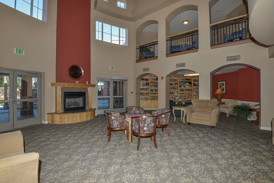 Spacious assisted living facility common area with high ceilings and large windows. The room features a fireplace with a red accent wall, a seating area with four patterned chairs around a small table, and additional armchairs and sofas. There are bookshelves filled with books visible through arched openings, and a balcony with railings overlooks the space. The carpet has a floral pattern, and there are exit signs above the doors.