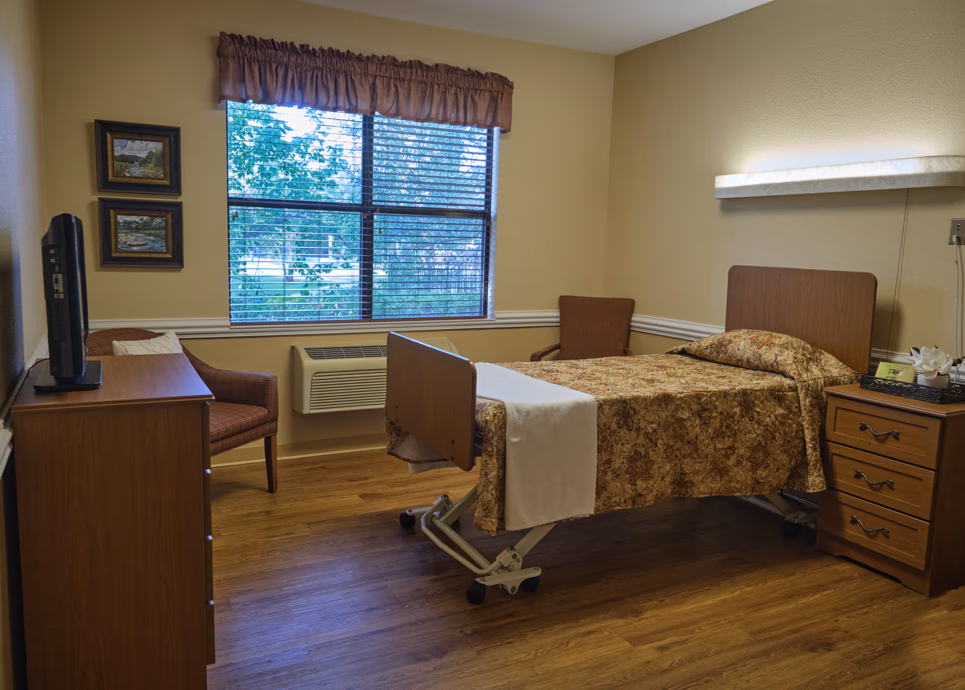 A furnished senior living bedroom with a single adjustable bed, bedside drawers, a TV on a dresser, chairs, and a window with blinds.