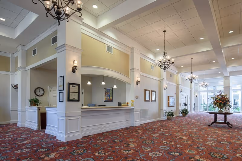 Bright, elegant reception lobby with a long white front desk, chandeliers, patterned red carpet, and a floral centerpiece near the entrance.