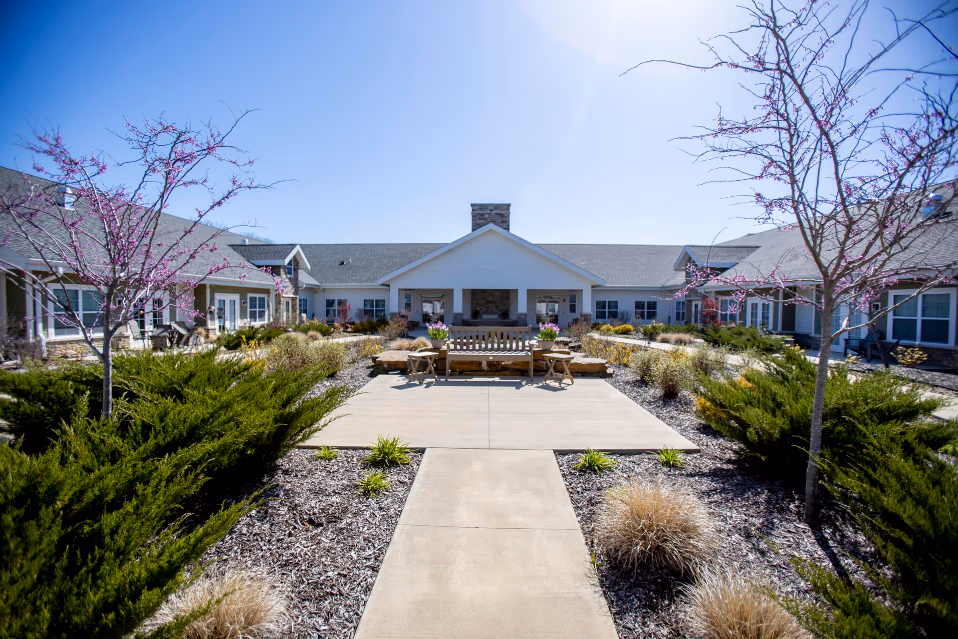 Outdoor courtyard area of a senior living facility with a concrete pathway leading to a wooden bench and small tables. The courtyard is landscaped with bushes, ornamental grasses, and trees with pink blossoms. The building surrounds the courtyard with multiple windows and a covered patio area with a stone fireplace.