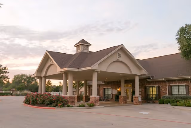 Exterior view of Legend Oaks Rehabilitation and Healthcare - Kyle building entrance with a covered drop-off area supported by columns, surrounded by landscaping and a driveway.