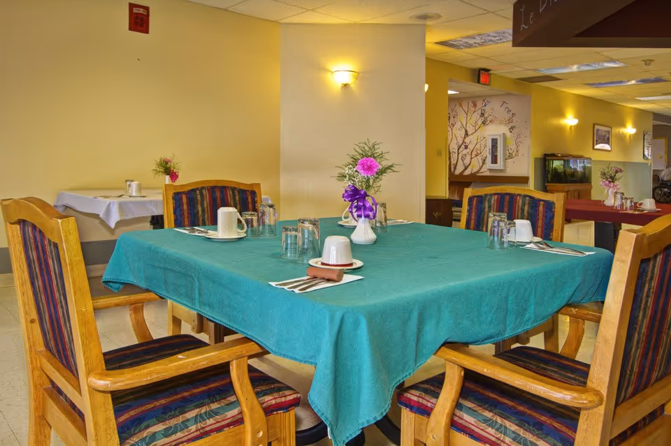 A dining area with a square table covered with a teal tablecloth. Four wooden chairs with striped cushions surround the table. The table is set with cups, glasses, napkins, and silverware, and a small vase with flowers is in the center. The room has yellow walls and additional tables and chairs in the background, along with wall decorations and a fish tank.
