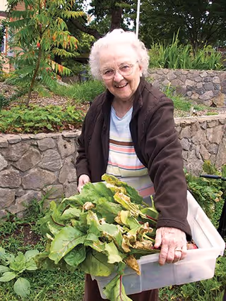 An elderly woman with white hair and glasses is smiling while holding a plastic container filled with freshly harvested leafy greens in a garden with stone walls and greenery in the background.