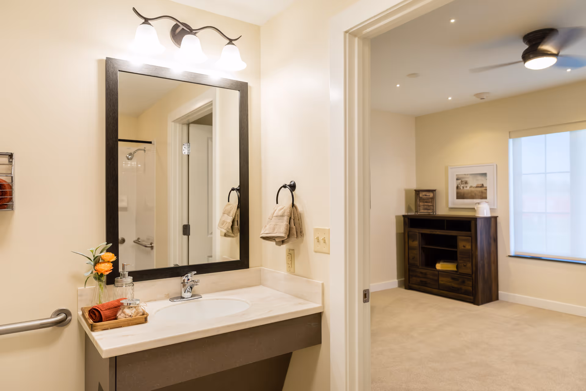 A bathroom sink area with a large rectangular mirror framed in dark wood, three overhead lights, a towel ring with a beige towel, and a small tray holding a flower vase, soap dispenser, and a rolled red towel. The bathroom opens into a carpeted room with a wooden cabinet, a framed picture on the wall, a window with a shade, and a ceiling fan with a light.