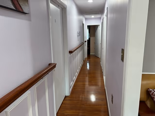 A narrow hallway with wooden flooring and light purple walls. There are white doors on both sides of the hallway, and wooden handrails run along the walls. The hallway is well-lit with ceiling lights, and a glimpse of a room with a brown couch is visible on the right side.