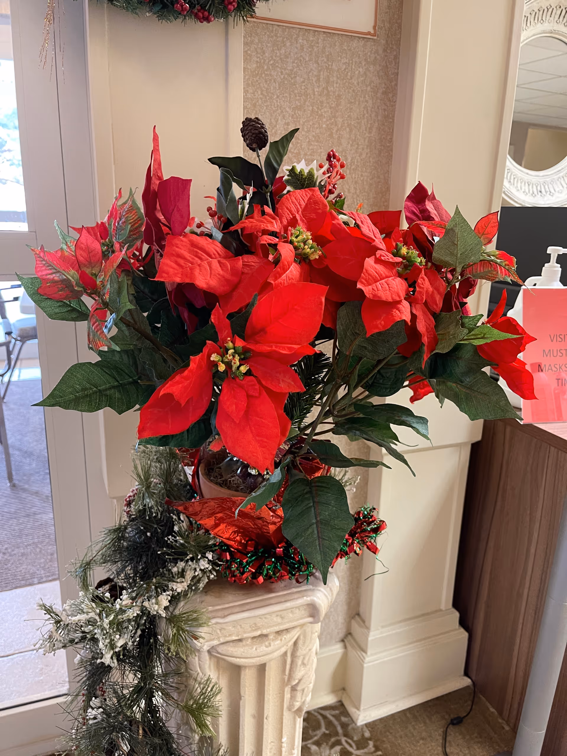 A festive holiday arrangement featuring bright red poinsettia flowers and green leaves placed on a decorative white pedestal. There is a garland with snow-like accents draped around the pedestal. In the background, part of a room with a mirror, a hand sanitizer dispenser, and a sign with red text is visible.